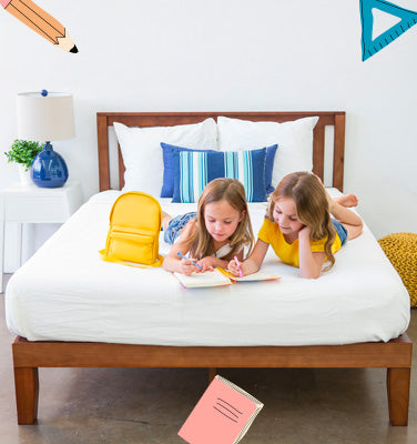 Two children lying on a bed with white bedding, blue pillows, and a yellow cushion.