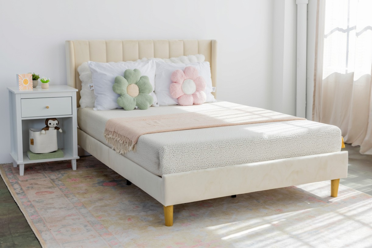 A light beige upholstered bed frame with a matching headboard and footboard, styled with white bedding, two flower-shaped pillows, and a pink throw blanket. The bed is shown in a bright bedroom setting next to a white nightstand.