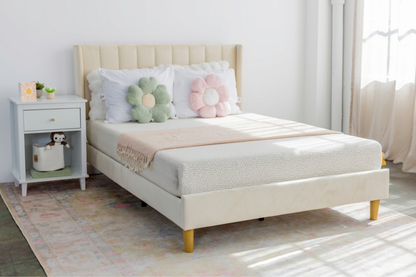 A light beige upholstered bed frame with a matching headboard and footboard, styled with white bedding, two flower-shaped pillows, and a pink throw blanket. The bed is shown in a bright bedroom setting next to a white nightstand.