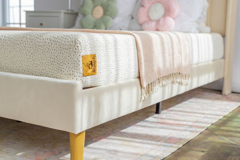 A close-up of a cream upholstered bed frame with a 5 little monkeys kids mattress, a light pink fringed throw blanket, and a yellow brand patch on the side, set in a softly lit bedroom with flower-shaped pillows and a striped rug.